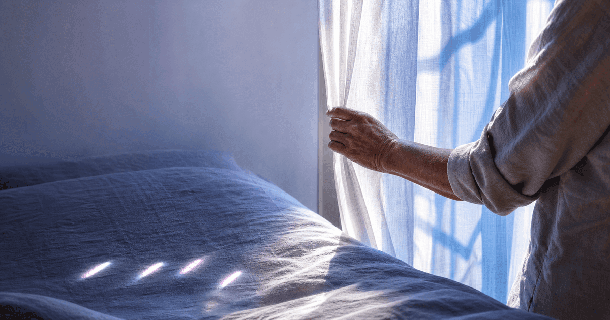 Older adult opens a sheer curtain at dawn beside four lavender light marks on textured bedding