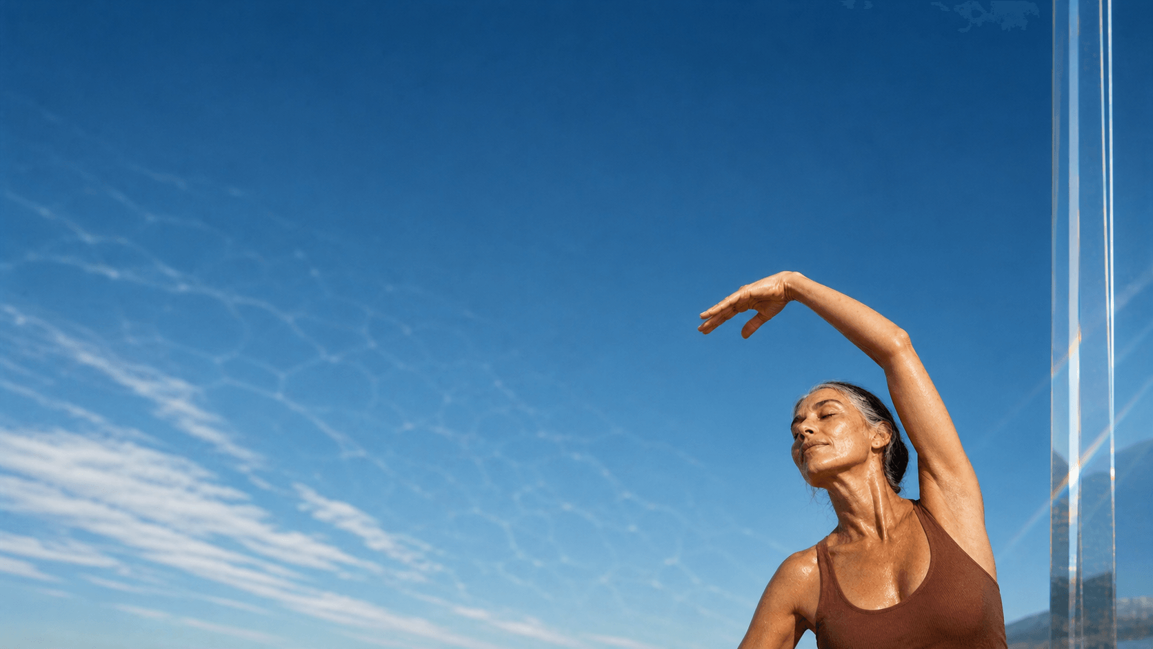 Older woman doing an overhead yoga stretch in hard sunlight beside refracted glass for cellular energy support.