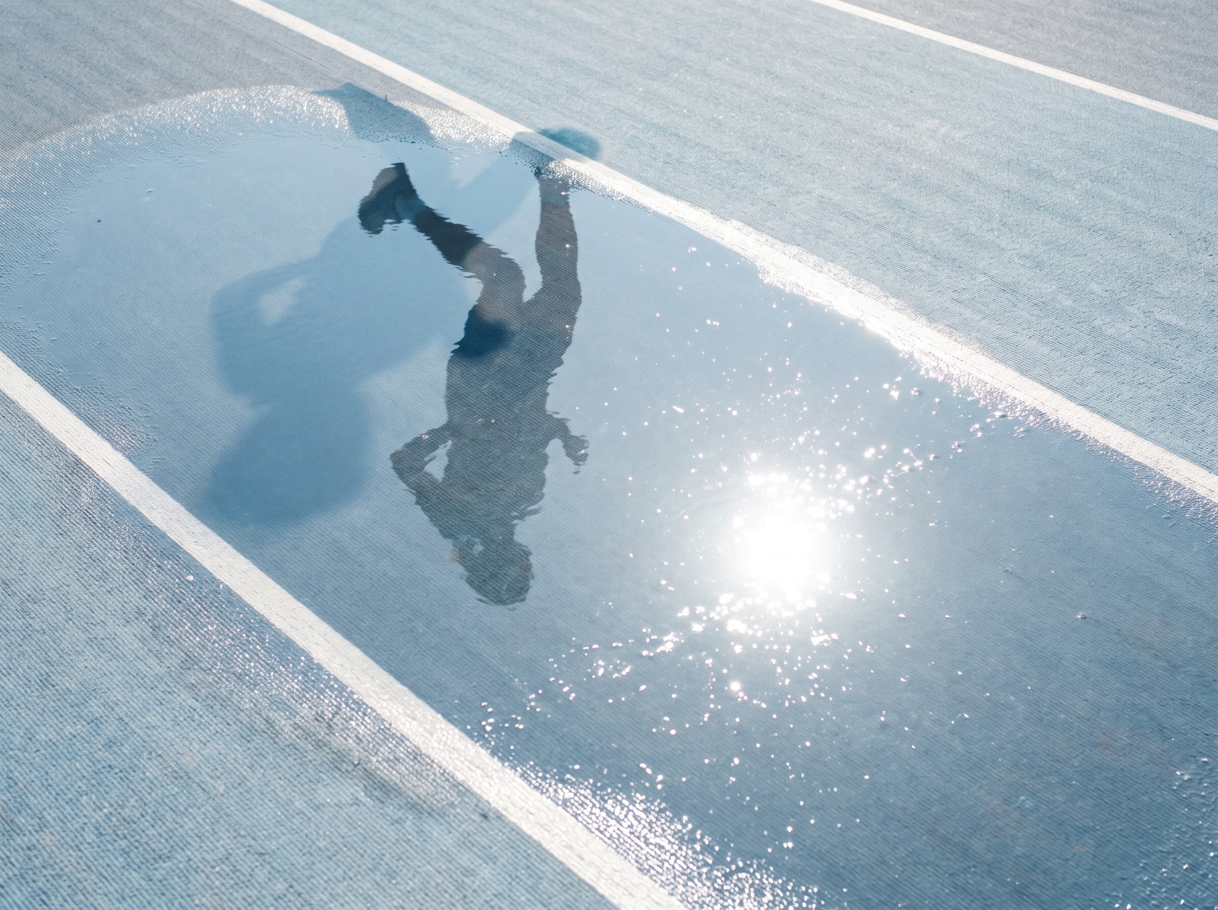 An athlete reflected in a bright puddle across a pale blue track.