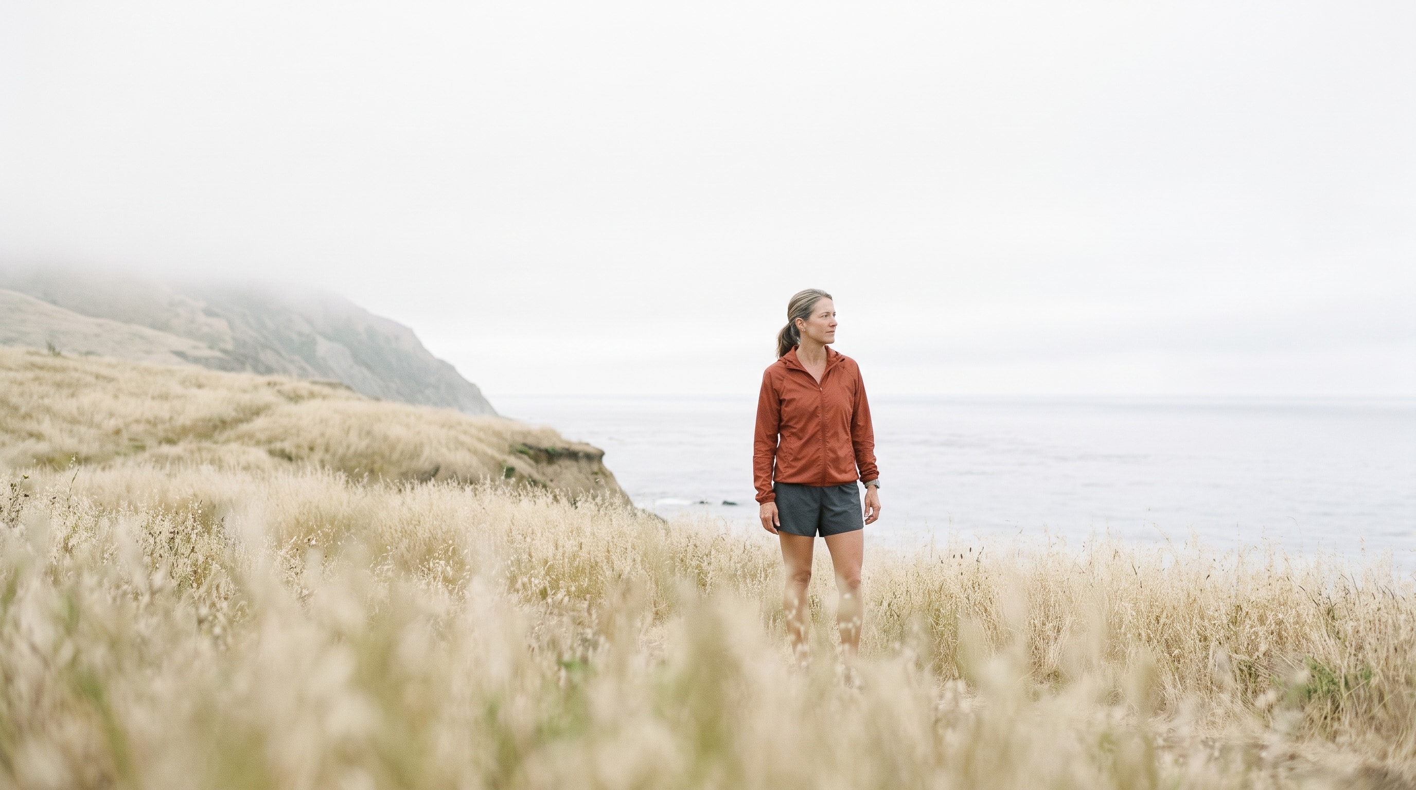 A runner stands in pale coastal grass beside a calm ocean.