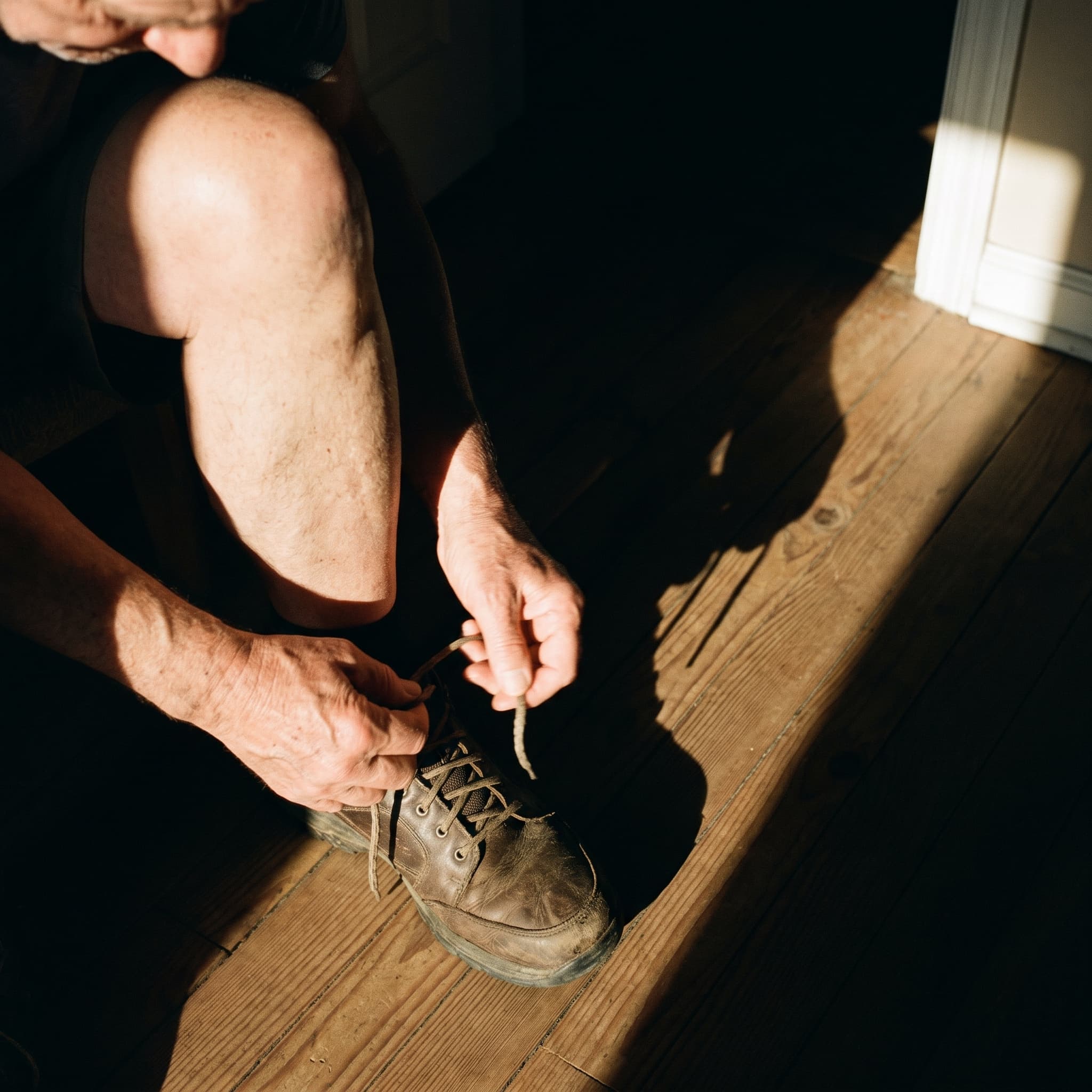Older man tying a worn walking shoe in a hard bar of doorway sunlight