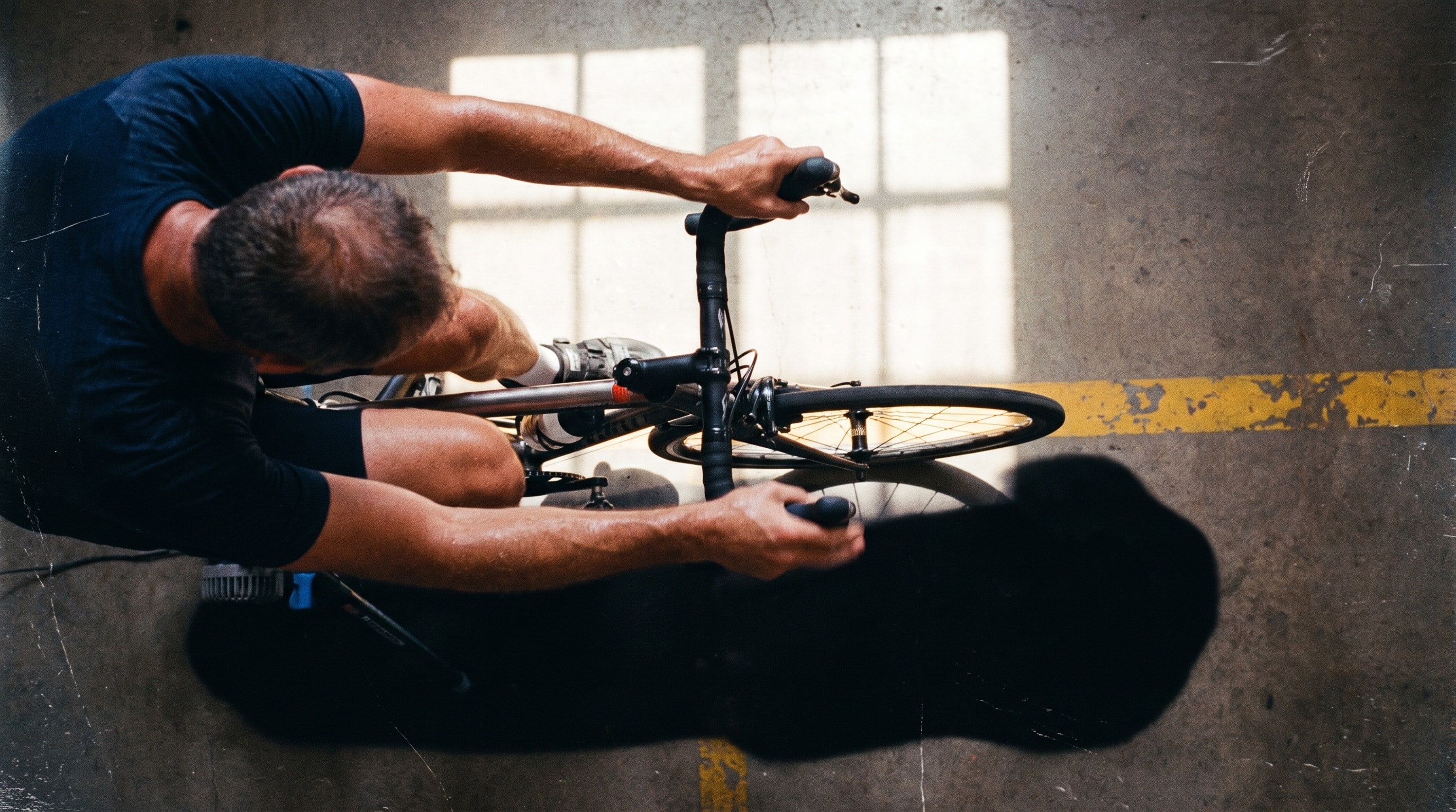 Cyclist gripping indoor training handlebars in a strip of hard sunlight.