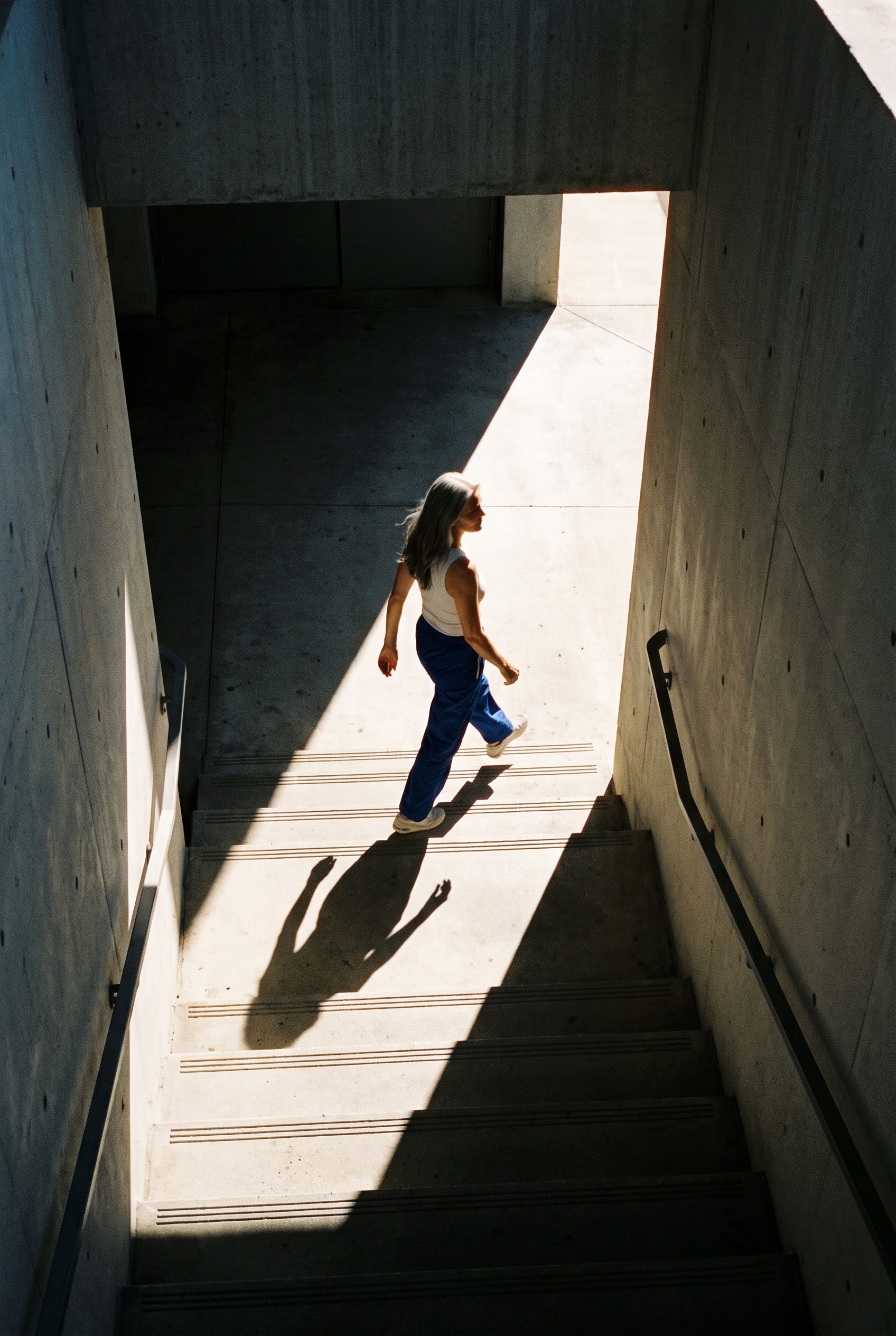 Woman crossing a shaft of light at the top of a concrete stairwell.