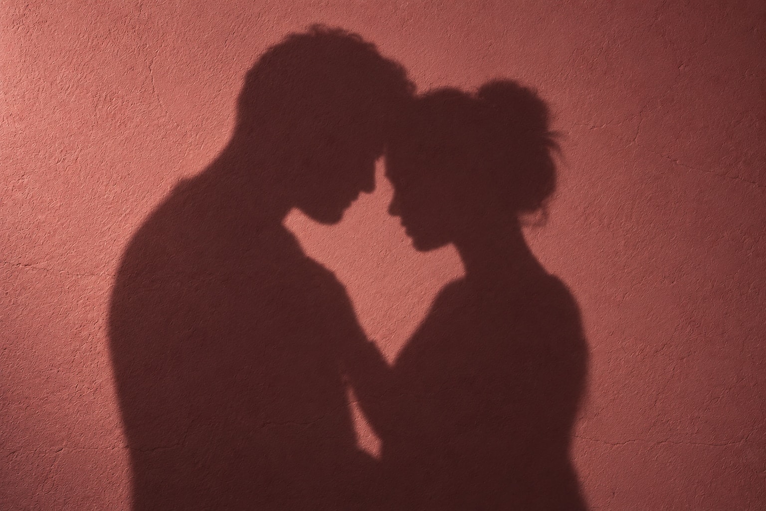 Shadows of two adults touching foreheads on a rose-colored plaster wall.