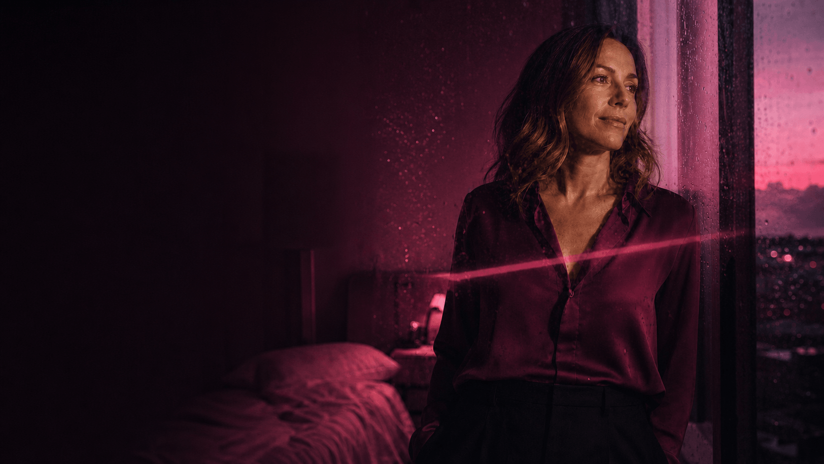 Woman in a dark berry blouse standing at a rain-spattered window in pink evening light.