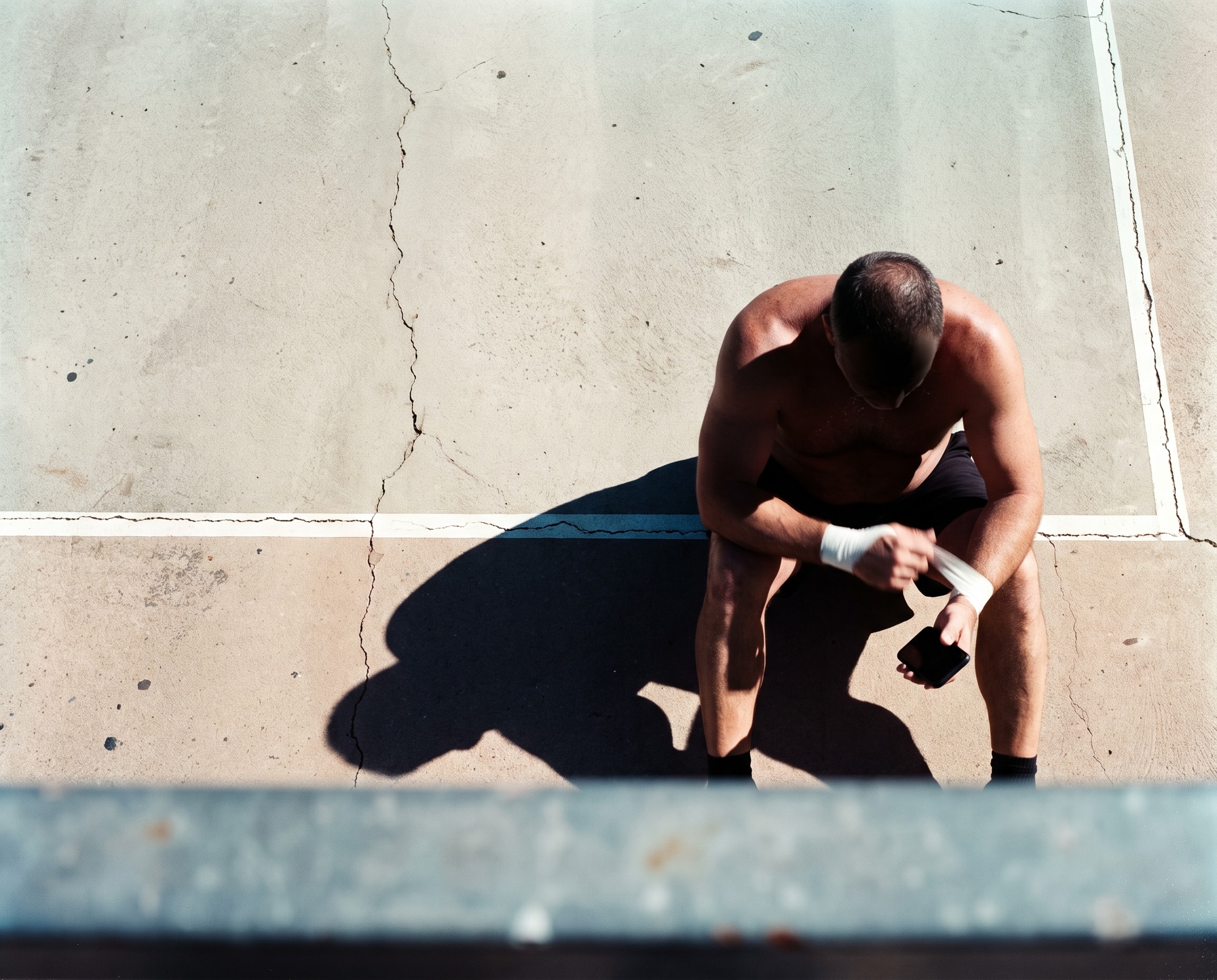 Athlete resting courtside in hard sunlight, taping a wrist and checking a phone.
