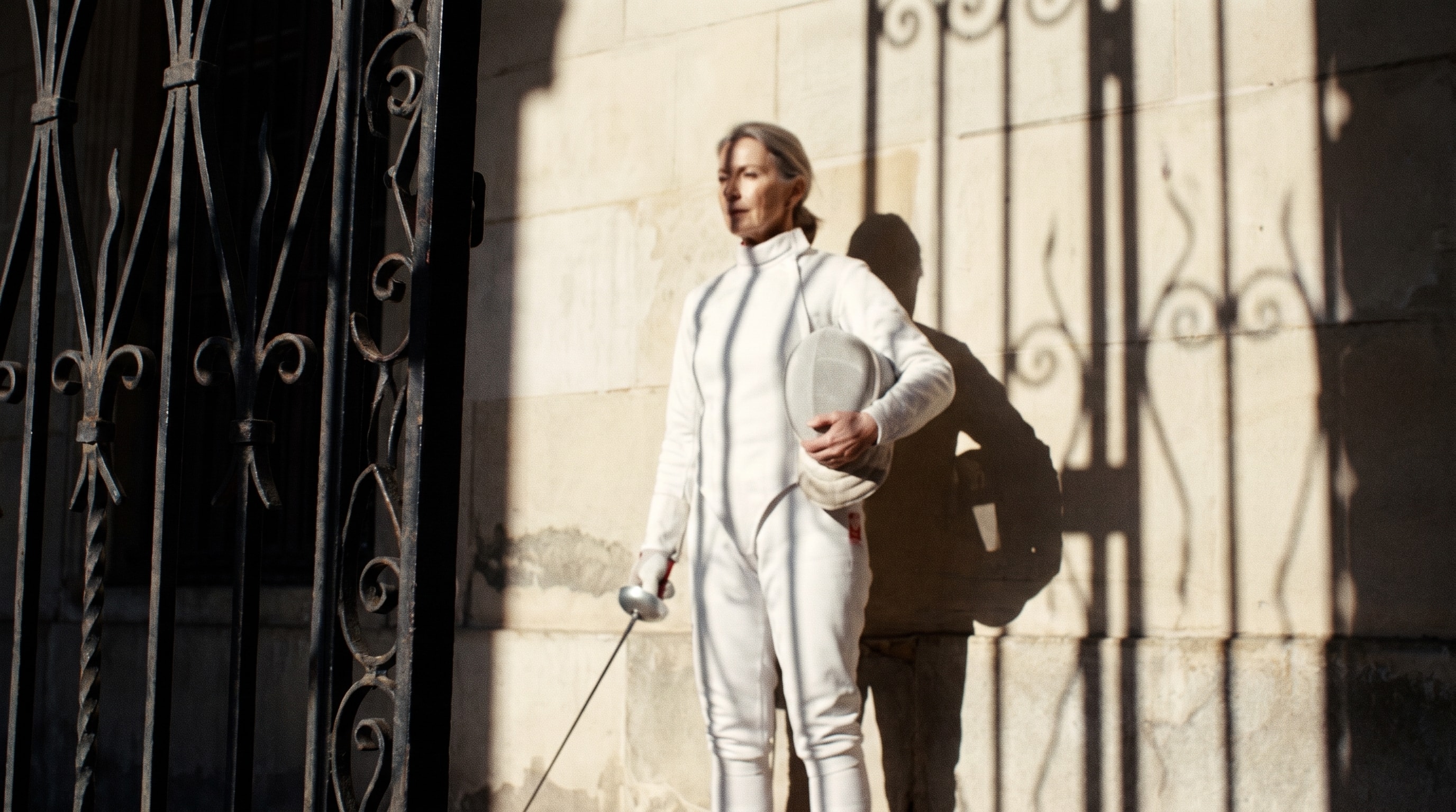 Fencer in white standing in low sun beside an iron gate.