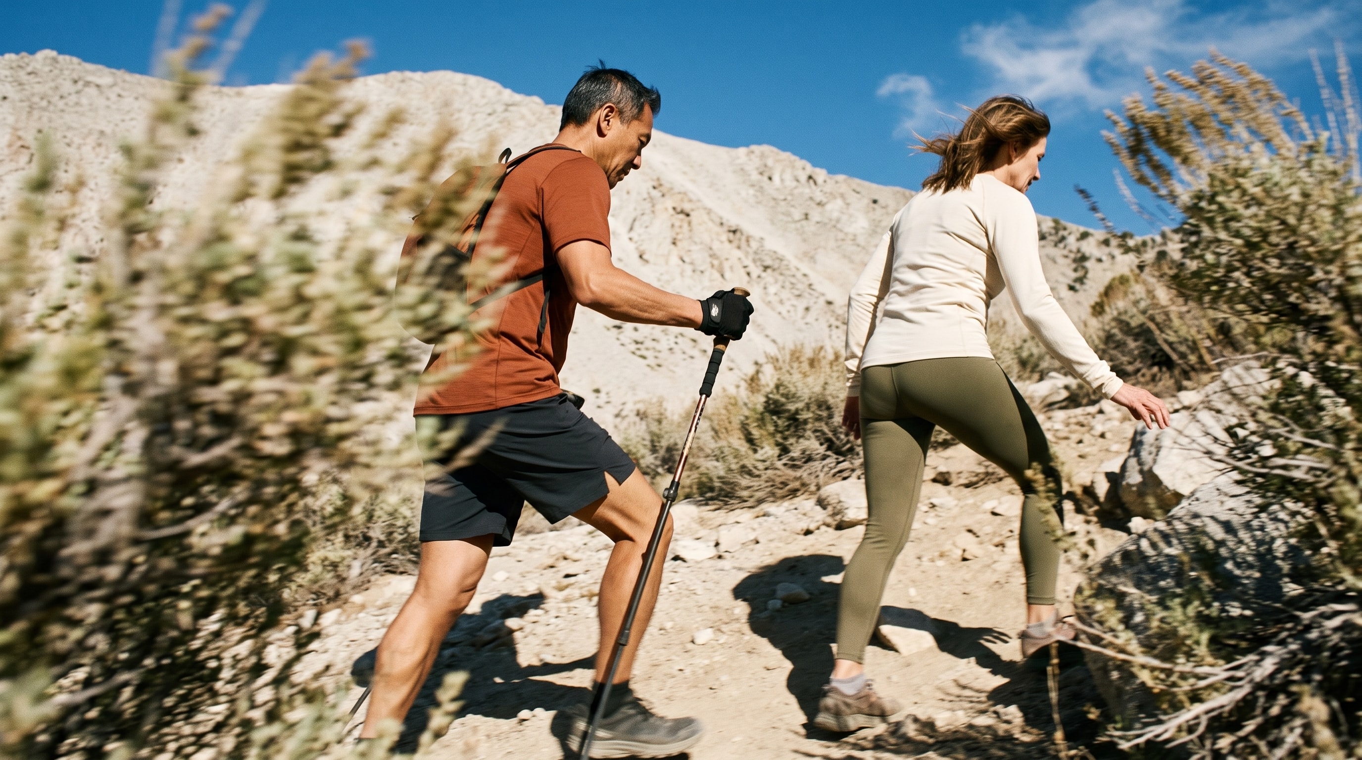 Two adults hike a bright alpine trail under a clear sky.