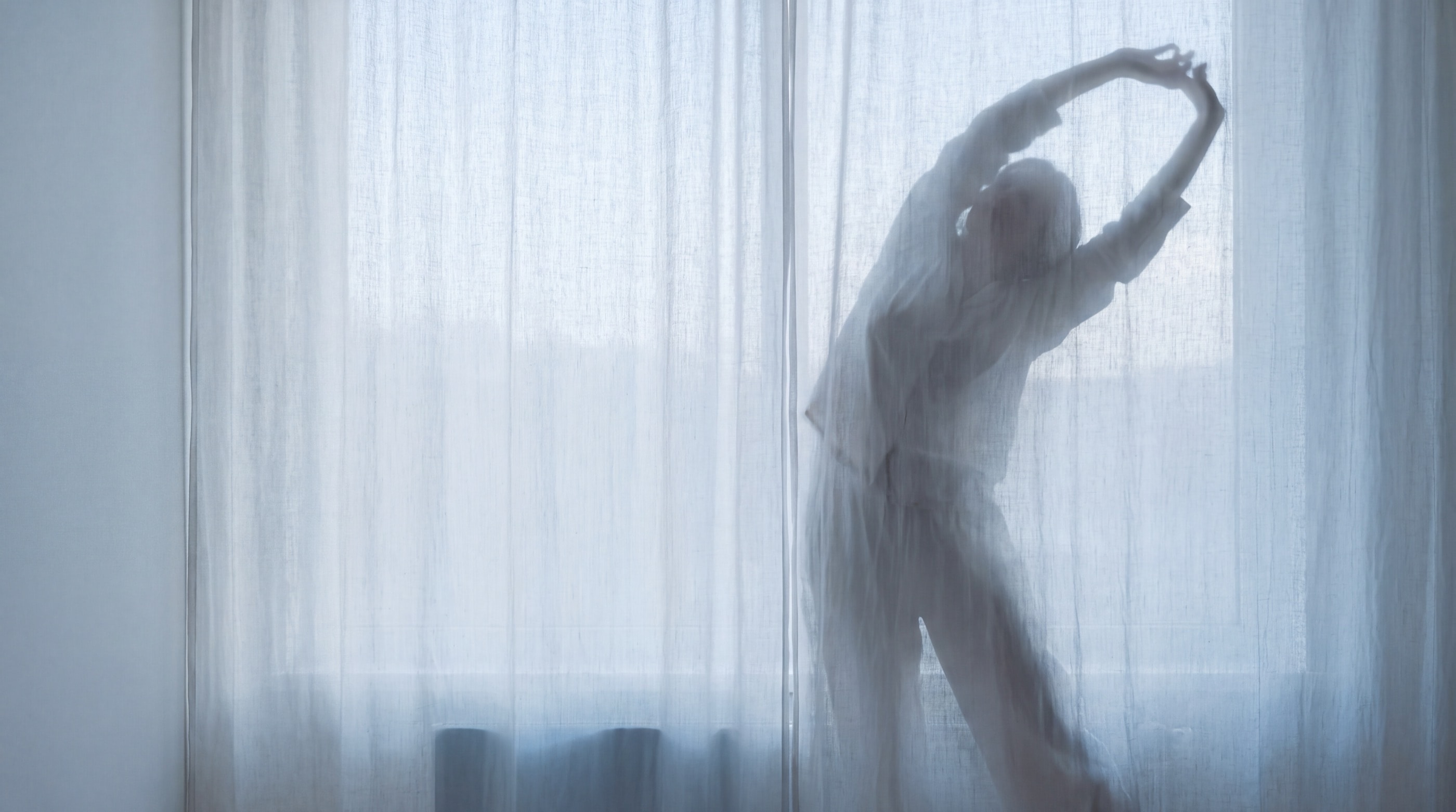 A person stretches in soft morning light behind a sheer curtain.